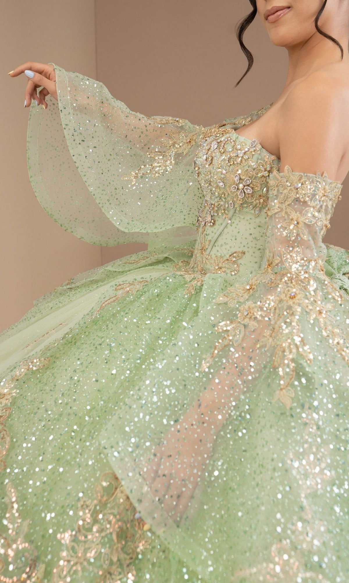 A person wearing the Quinceanera Dress 8575JQ By Cinderella Coutureโa sparkly, light green embellished ball gown with gold embroidery and sheer, flared sleeves; only the upper body is visible.